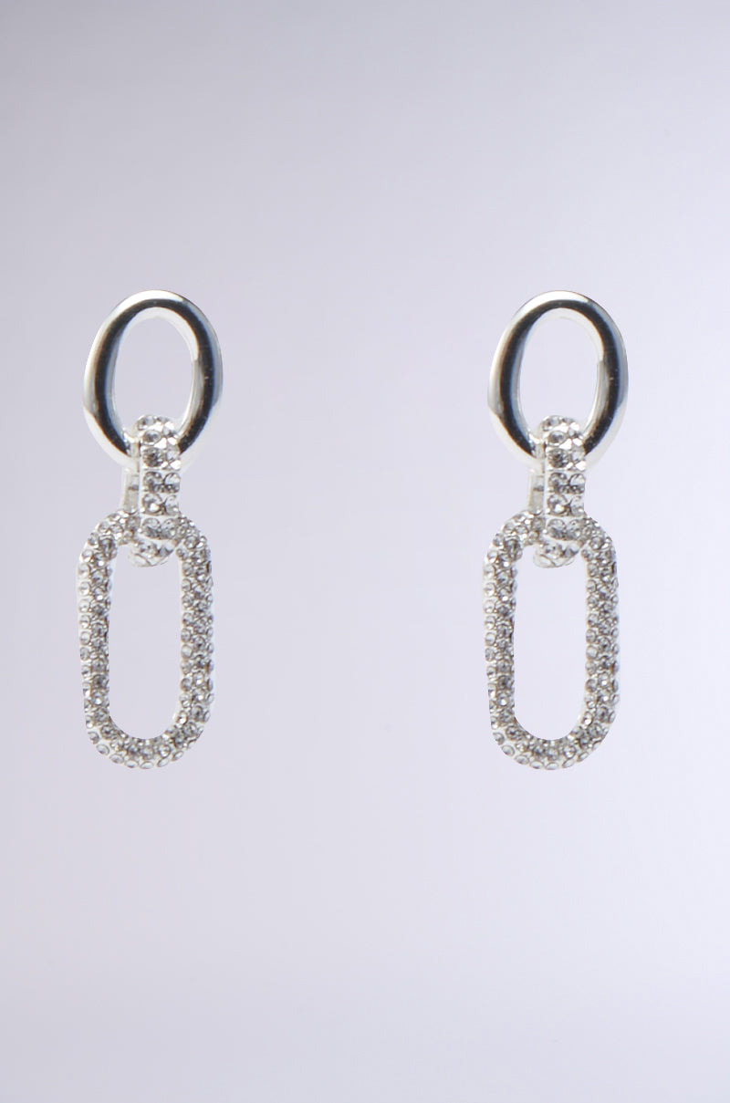 The PAVE DANGLE EARRING by AKIRA Label showcases silver earrings with an oval hoop and a larger elongated rectangular loop, embellished with sparkling cubic zirconia stones. The background features a gradient from light gray to white. 0