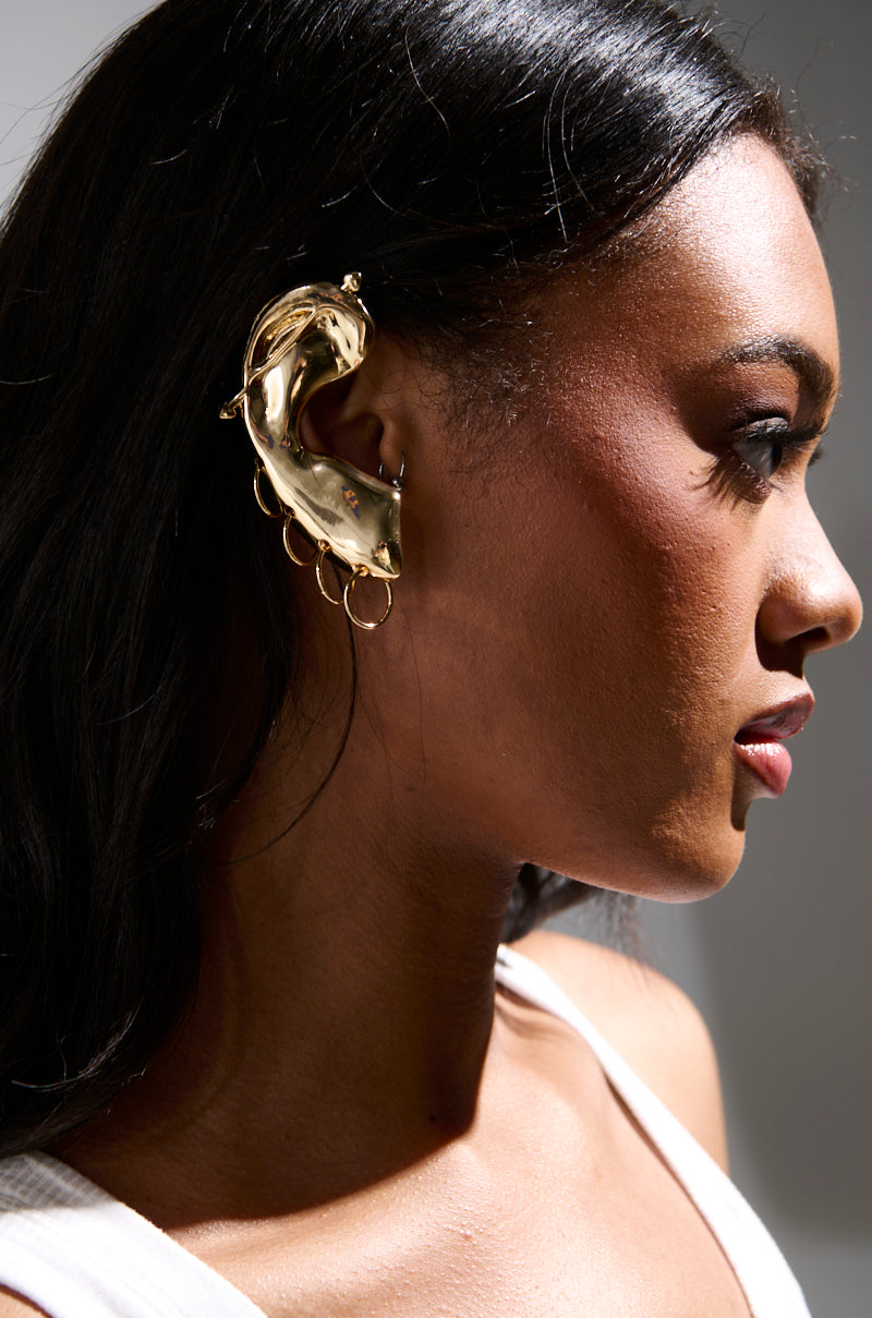 In profile against a soft gray background, a person with long, dark hair wears the HEARD YOU EAR CUFF by AKIRA Label. Adorned with decorative rings, this striking faux gold accessory beautifully complements their white top as they gaze to the right.
