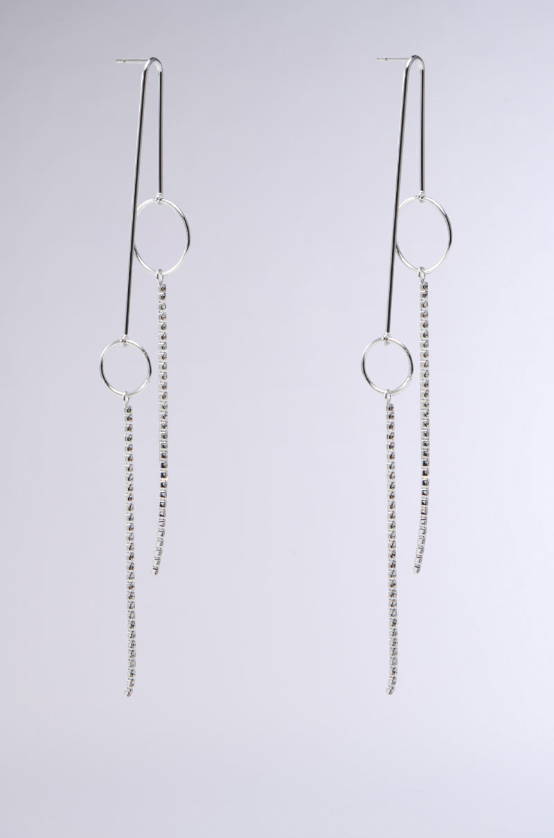 The RUNNING IN CIRCLES EARRING by the AKIRA Label features two elegant silver earrings with a thin vertical bar, circular loop, and cascading delicate chain strands, displayed on a plain background for added sophistication.