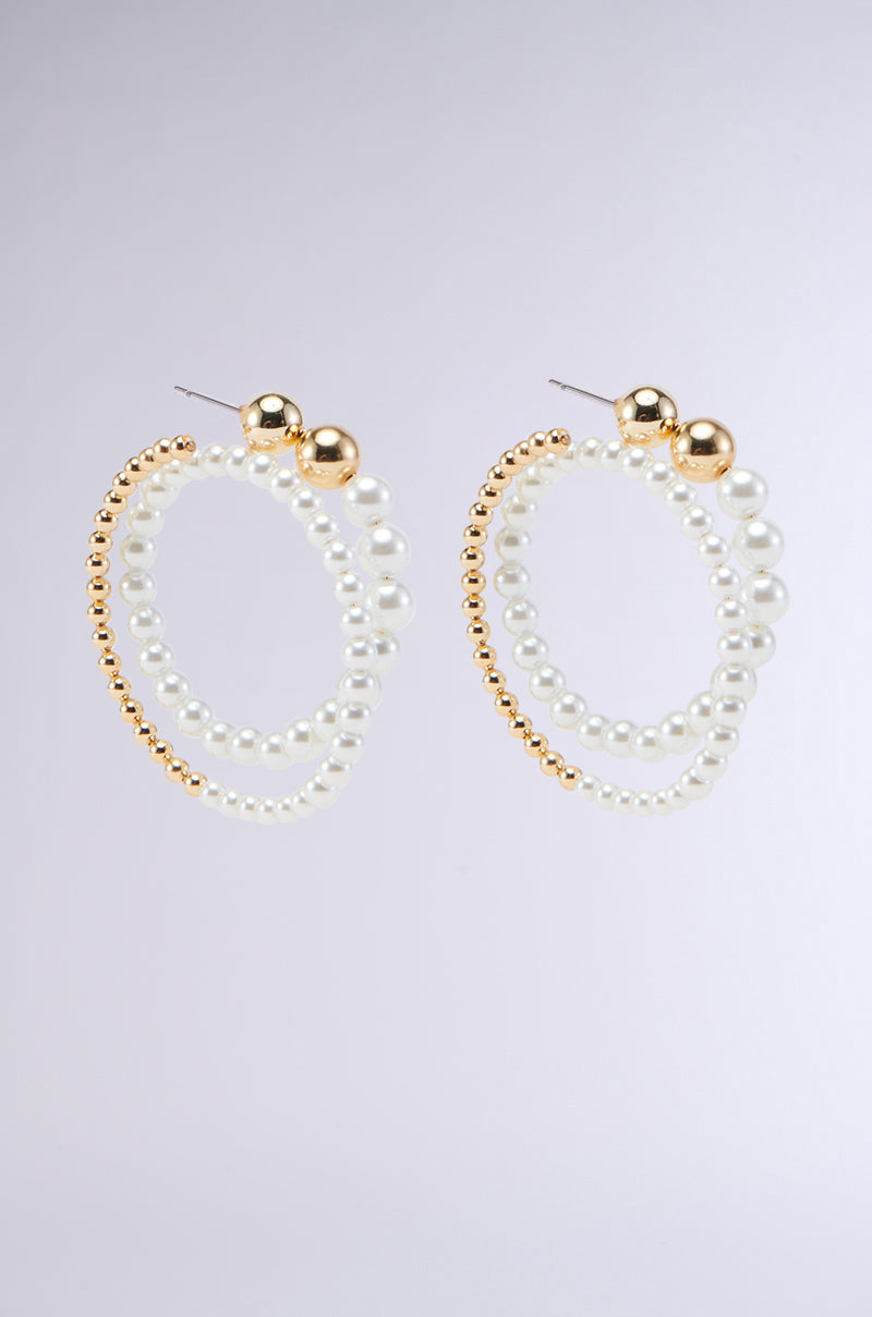 The SPIRALING EARRING features a pair of dangling hoop earrings with two interlocking loops; one loop crafted from small gold beads and the other combining gold and white pearl-like beads. These earrings are finished with classic post-back closures, adding to their sleek and elegant appearance against a neutral background.