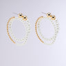 The SPIRALING EARRING features a pair of dangling hoop earrings with two interlocking loops; one loop crafted from small gold beads and the other combining gold and white pearl-like beads. These earrings are finished with classic post-back closures, adding to their sleek and elegant appearance against a neutral background.