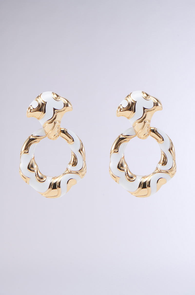 The VENETIAN LOVE EARRING showcases an exquisite pair of gold and white enamel earrings adorned with a unique wavy, twisted design. These interlocked dangle hoops boast an open circular shape and a decorative top piece, offering an elegant and distinctive appearance, set against a plain gray background.