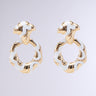 The VENETIAN LOVE EARRING showcases an exquisite pair of gold and white enamel earrings adorned with a unique wavy, twisted design. These interlocked dangle hoops boast an open circular shape and a decorative top piece, offering an elegant and distinctive appearance, set against a plain gray background.