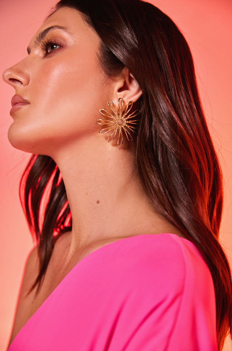 A woman with long brown hair wears striking IN BLOOM GOLD EARRINGS and a hot pink off-the-shoulder top, standing against a pink-red gradient background with dewy makeup highlighting her glowing skin.