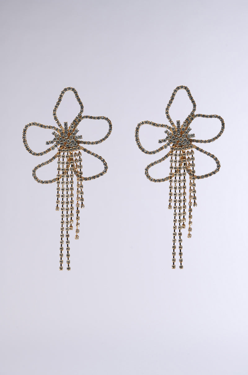 The "IN THE GARDEN EARRING" features gold statement earrings with flower-shaped silhouettes, intricate loops, and rhinestone-encrusted petals. Dangling chains resemble a floral stamen against a light grey background.