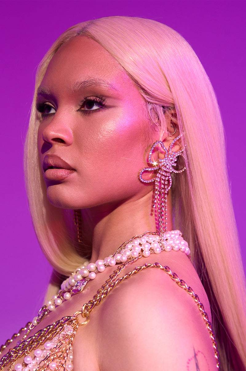 A person with long, straight platinum blonde hair poses against a purple background, wearing the IN THE GARDEN EARRING—large, flower-shaped statement earrings—along with layered pearl and gold chain necklaces.