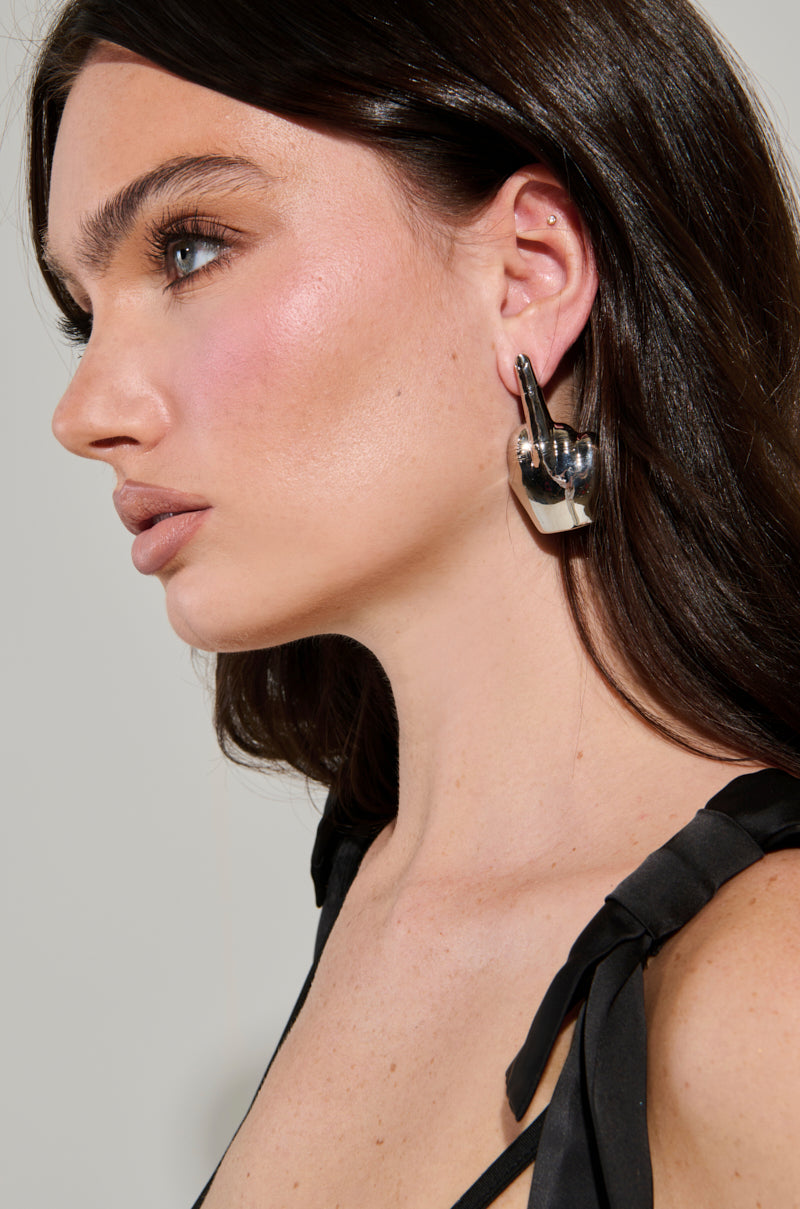 A woman with long dark hair and MY MOOD EARRING, a bold metallic statement-maker, gazes to the side. Sporting a subtle pink blush and natural makeup, she wears a black outfit with ribbon straps. Soft, neutral tones in the background complement her striking appearance. 1