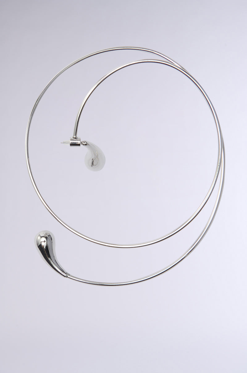 The WRAP ME AROUND EAR CUFF is a stylish, metallic accessory with a spiral hoop design. It has a curved metal structure resembling a modern lamp, featuring an abstract bulbous end with faux silver finish, set against a plain white background.