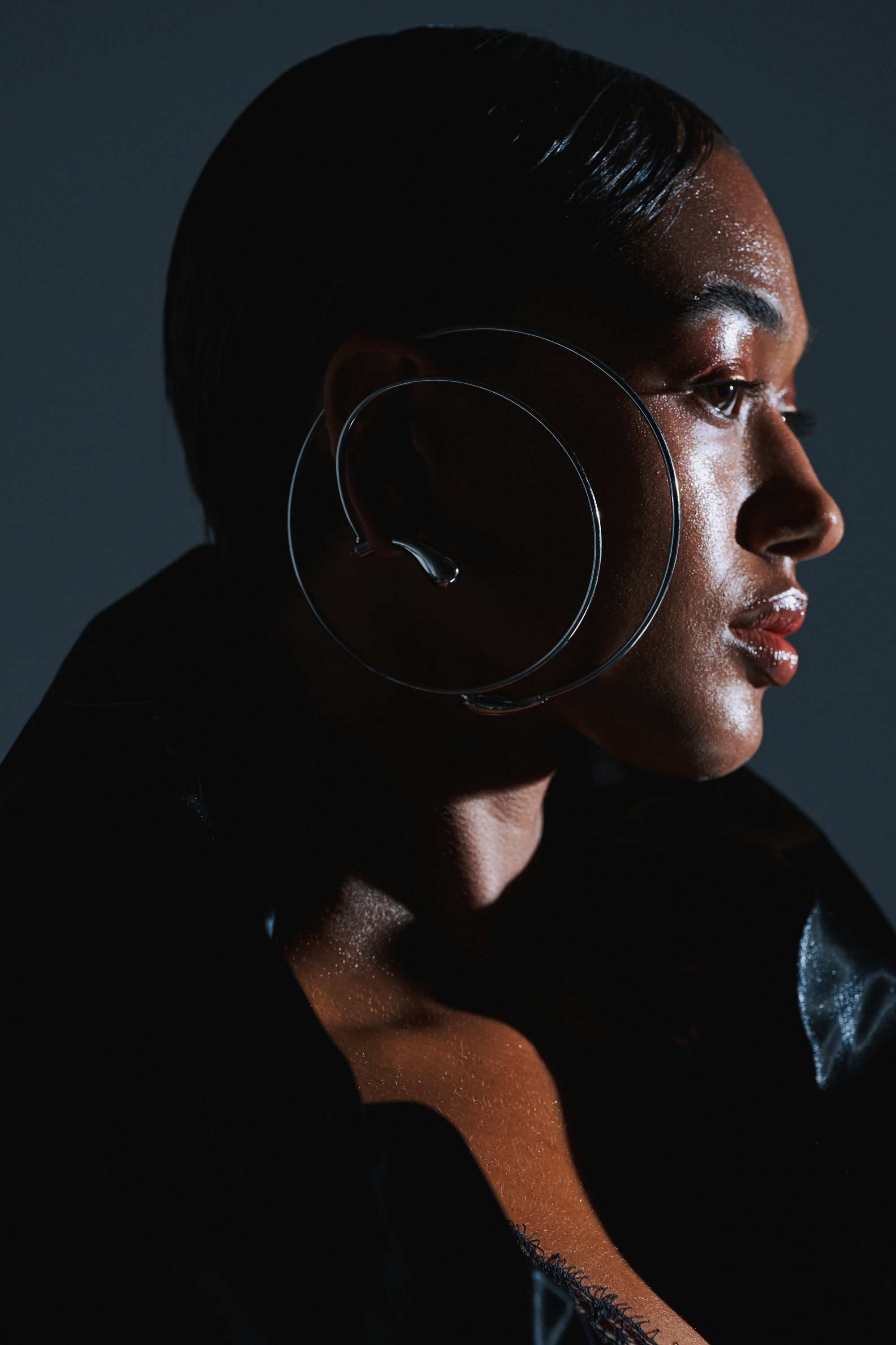 A person with glowing skin and slicked-back hair is shown in profile, wearing the WRAP ME AROUND EAR CUFF and a black textured outfit against a dark background.