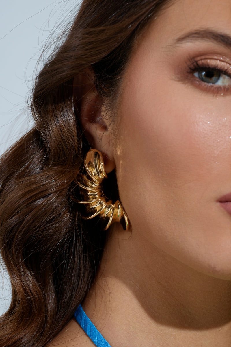 A woman's face is close-up, showcasing her BELLA NOCHE GOLD EARRING with a wavy design. Her long, wavy brown hair complements the glittery eye makeup and bright blue strap on her shoulder against a light, neutral background. 1