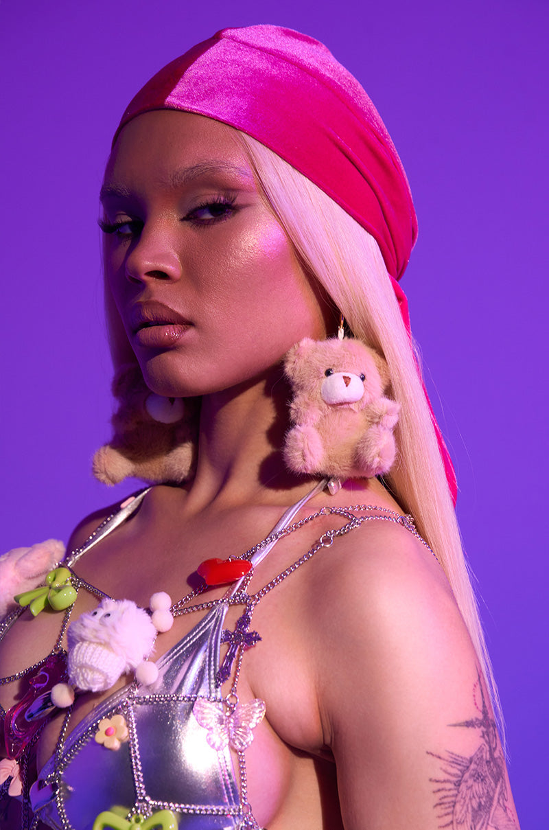 Wearing long blonde hair and bold makeup, they showcase BEAR WITH ME EARRING, a shiny pink headscarf, metallic top, toy-adorned jewelry, and a visible tattoo against a vibrant purple background.