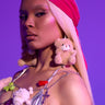 Wearing long blonde hair and bold makeup, they showcase BEAR WITH ME EARRING, a shiny pink headscarf, metallic top, toy-adorned jewelry, and a visible tattoo against a vibrant purple background.