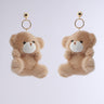 The BEAR WITH ME EARRING features adorable plush teddy bears with soft beige fur, white snouts, and tiny eyes. These chic accessories are attached to gold ball studs with delicate gold rings and set against a simple pale gray background.