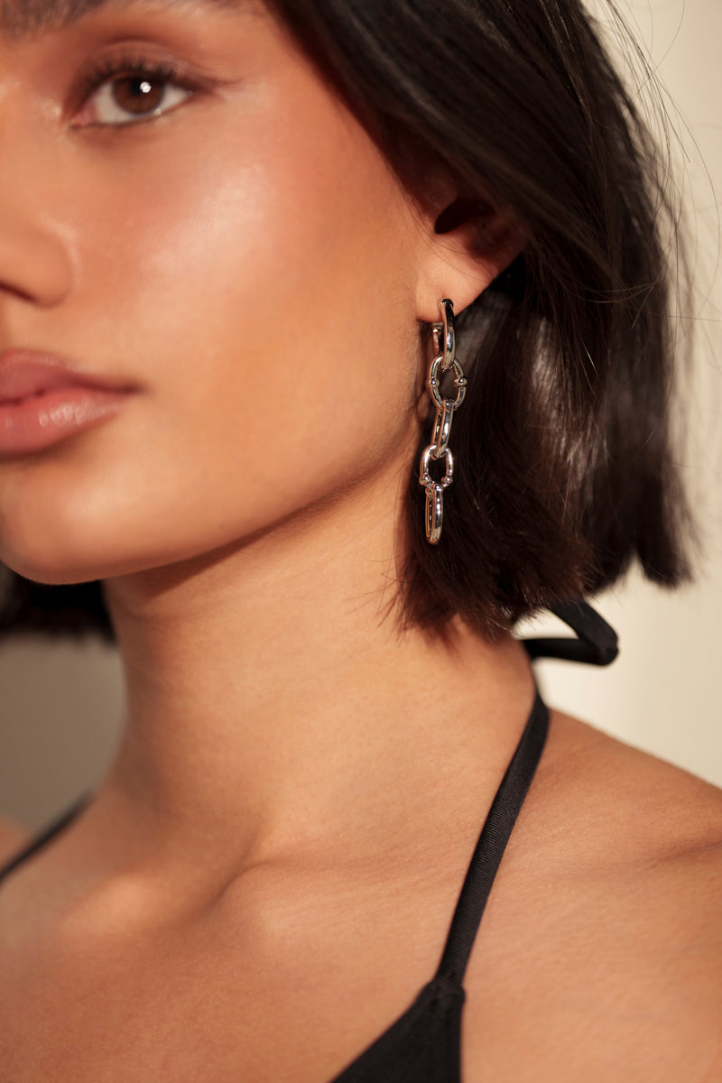 A close-up of a woman with short dark hair wearing the MOD GAL EARRING and a black halter top, illuminated by warm natural light. Her smooth skin is visible, with only part of her face shown.
