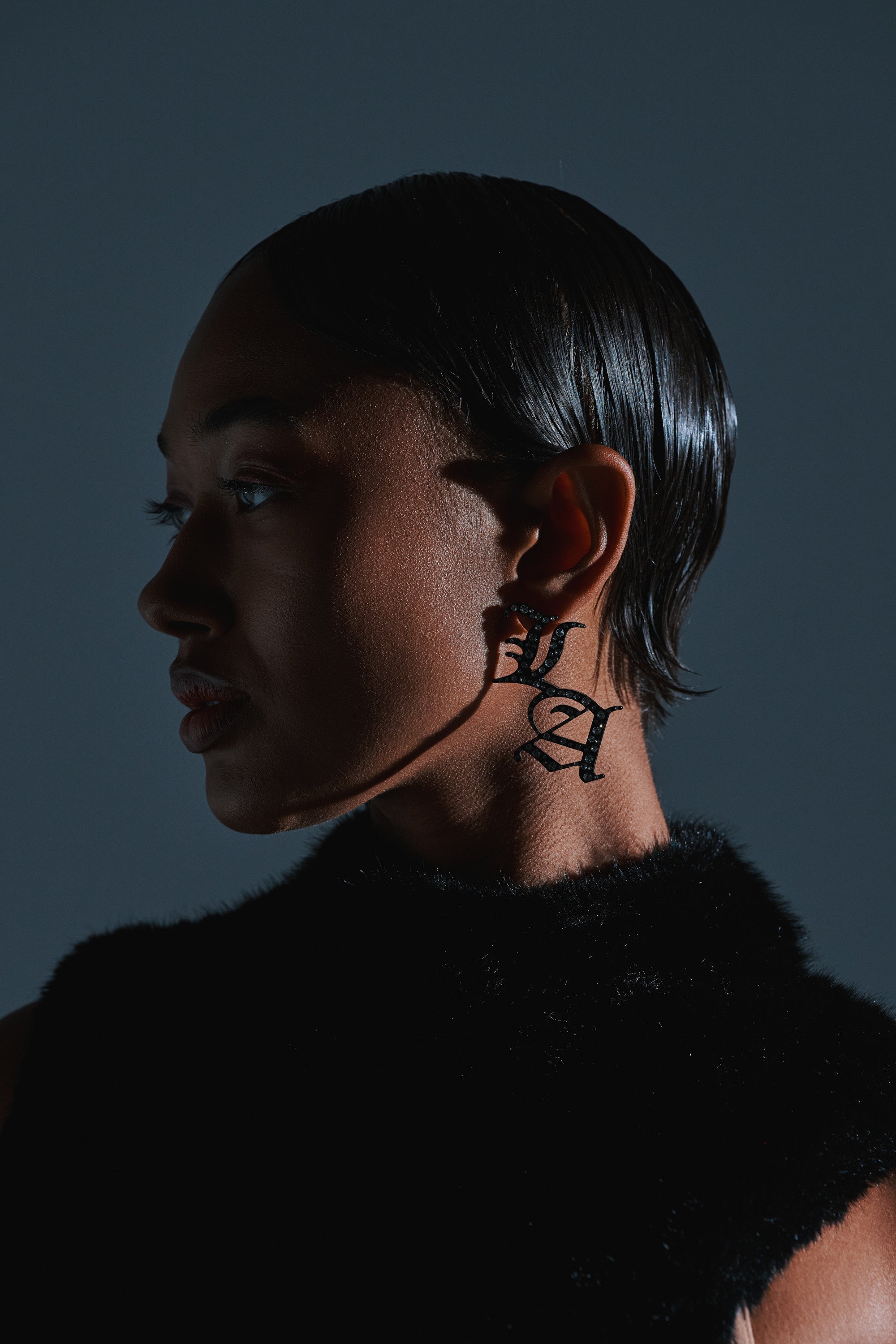 A person with slicked-back hair wears a black fuzzy top and WRITE ABOUT ME EARRINGS, facing left. On their neck, a shadow forms bold "L" and "A" letters. The background is dark gray. 1