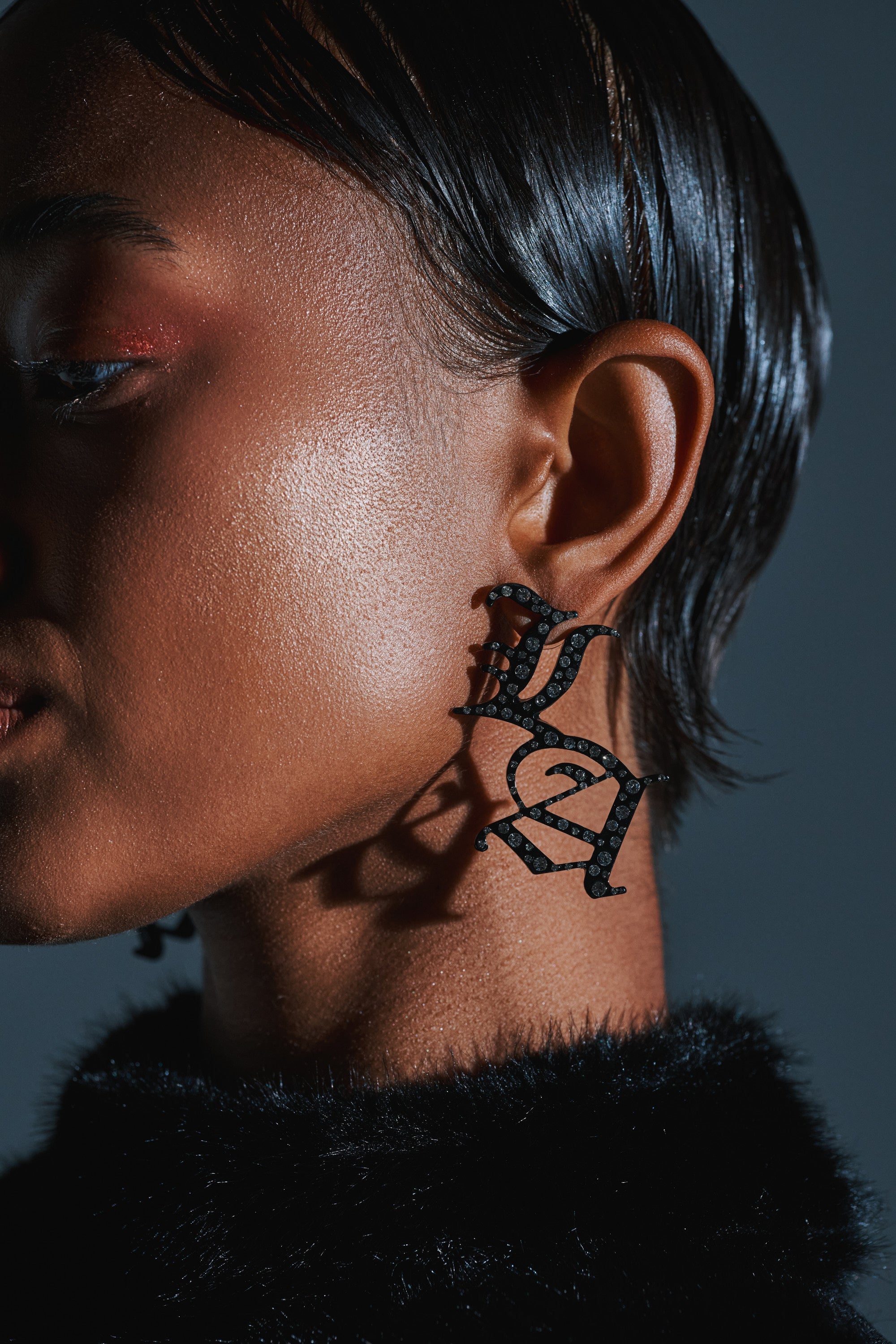 Close-up of a person with smooth, dark skin and sleek, wet-look hair wearing WRITE ABOUT ME EARRINGS—large black jewelry in old-English font—and a black fuzzy garment, set against a dark background. 3