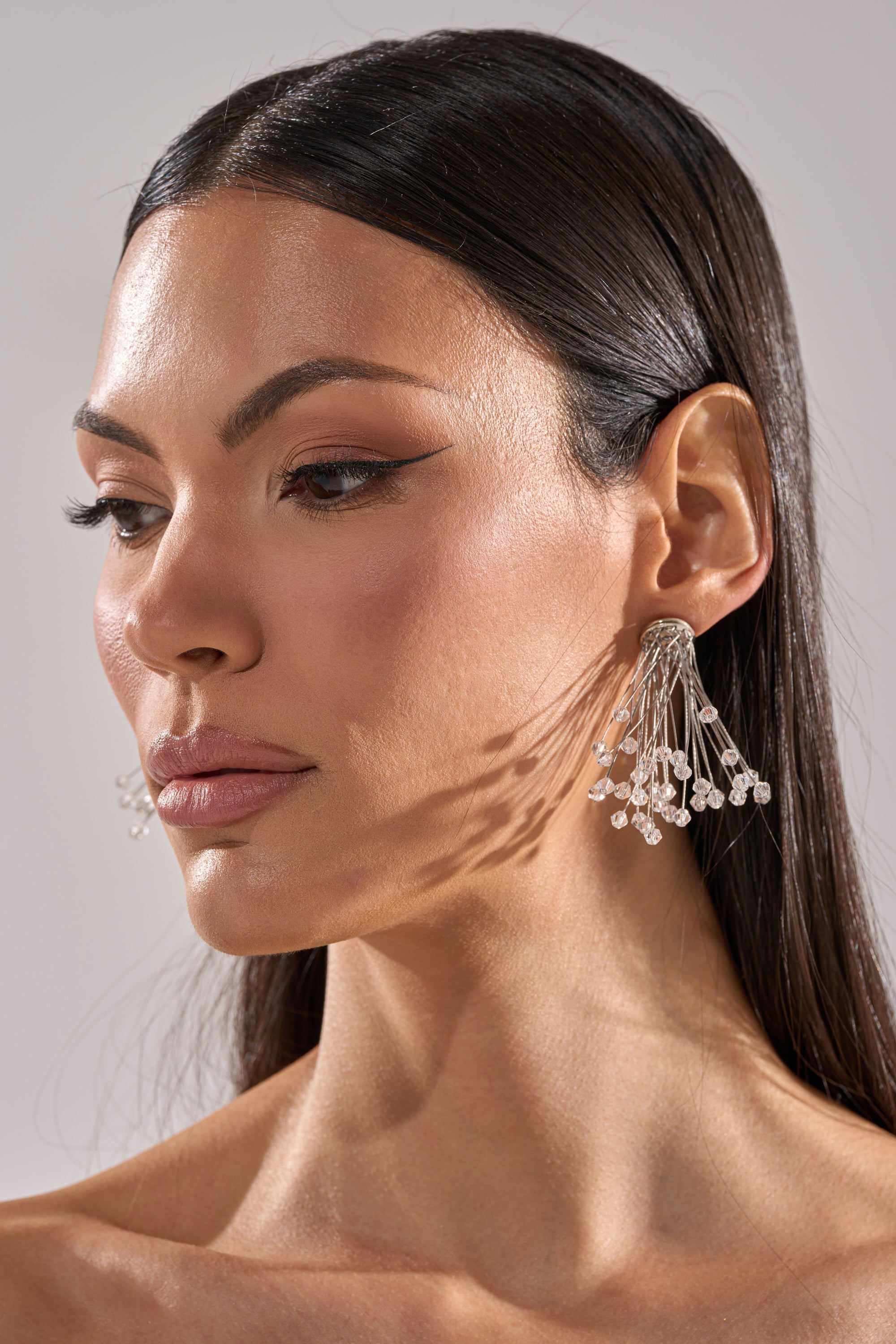 A woman with long, straight dark hair wears the IN PARIS EARRING—oversized earrings featuring multiple thin strands with small beads—casting shadows on her cheek. She has natural makeup and a neutral expression. 0