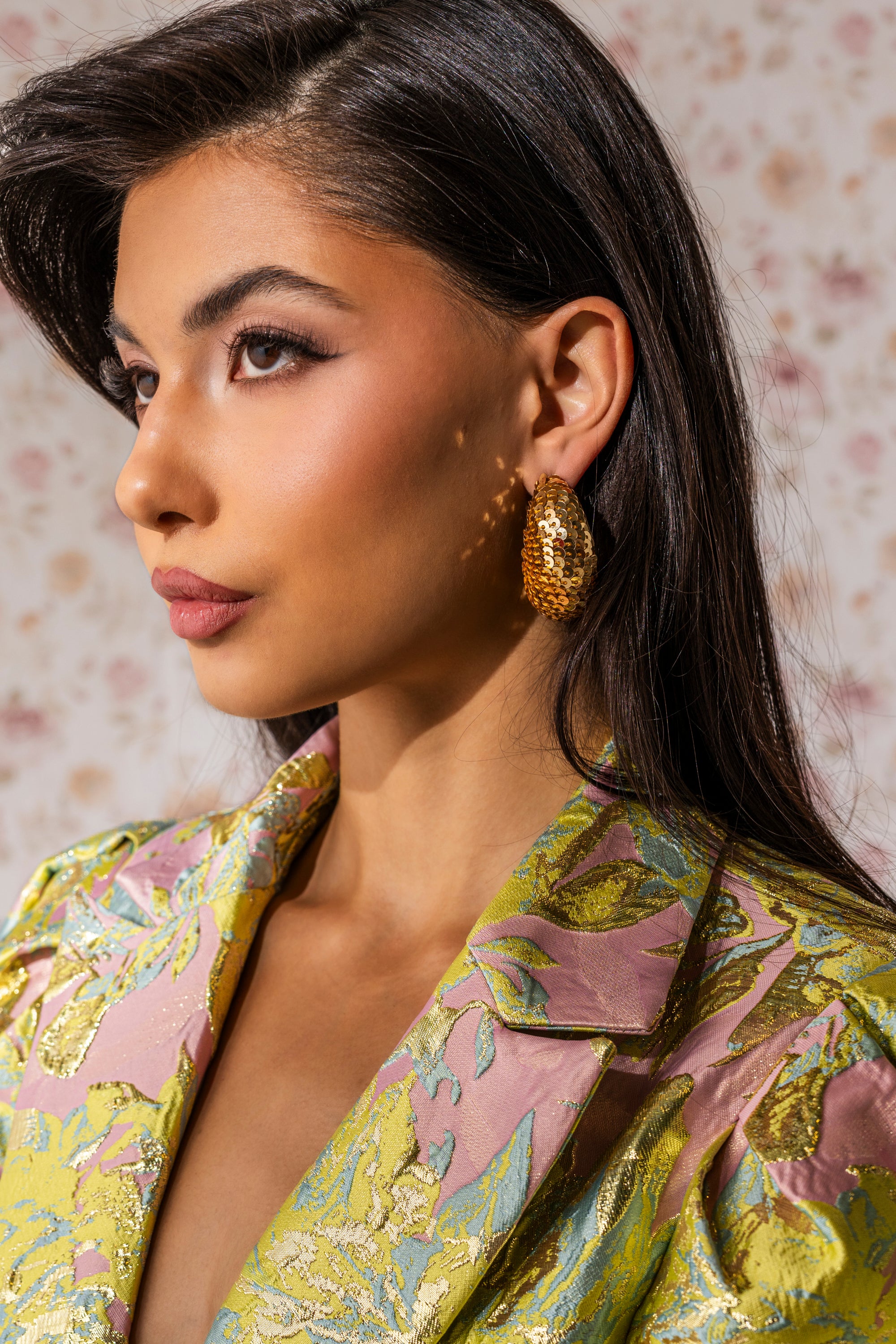 A woman with long dark hair wears a floral pastel blazer and the GIMME THE GLITTER HOOP EARRING, posing in profile against a soft floral background. 0
