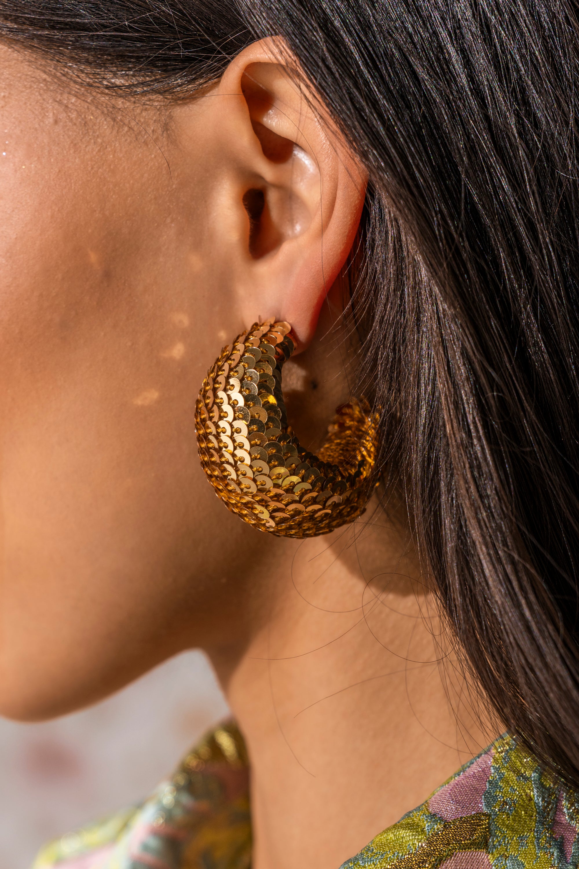 Close-up of a person's ear adorned with the GIMME THE GLITTER HOOP EARRING—large gold sequin hoops. Their dark hair is tucked behind the ear, paired with green and gold patterned fabric for a true disco glam style. 1