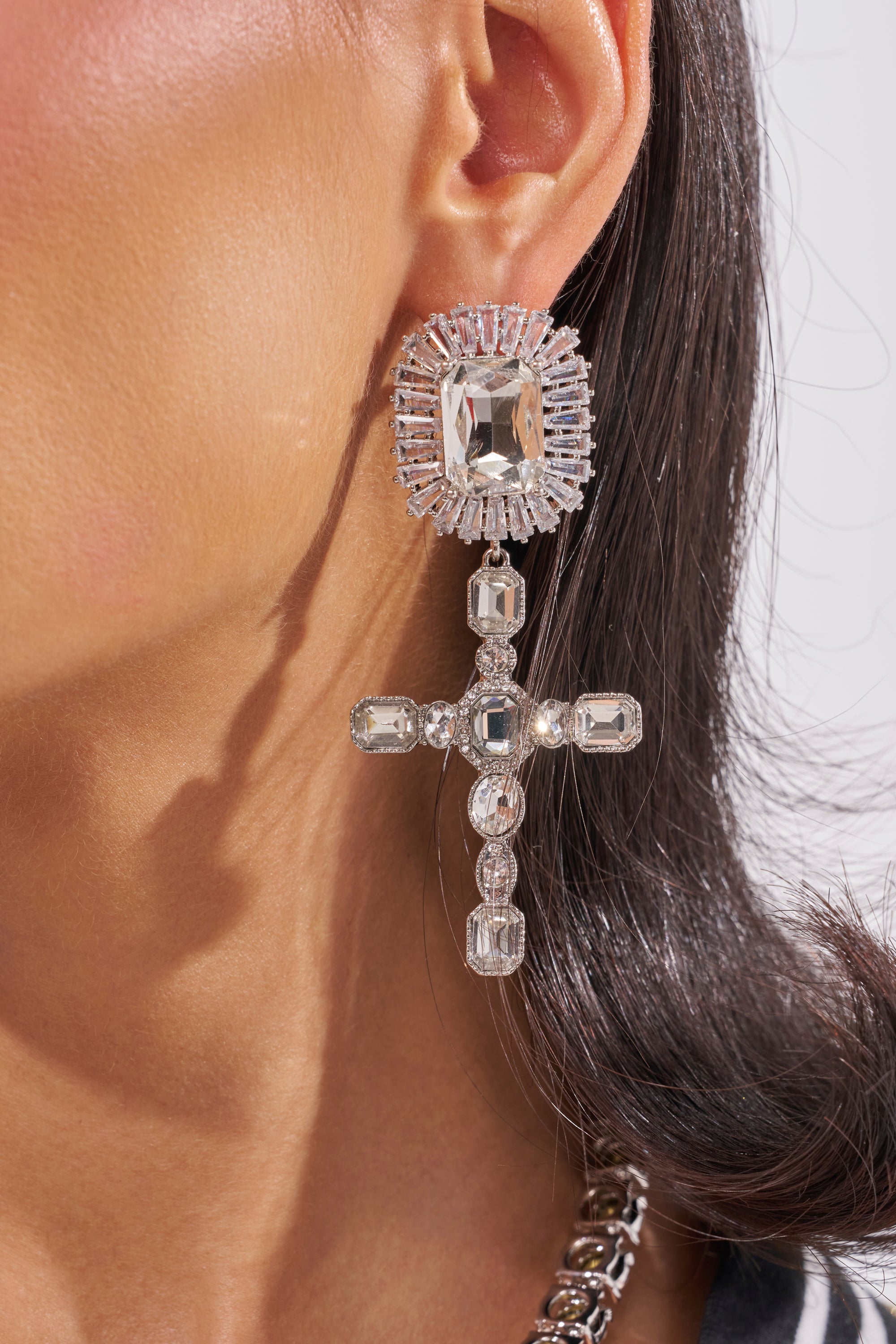Close-up of a person wearing the CROSS YA HEART EARRING: bold statement earrings featuring a large square gem at the top and a dangling cross shape made of clear crystals, paired with sleek dark hair for a striking look. 1