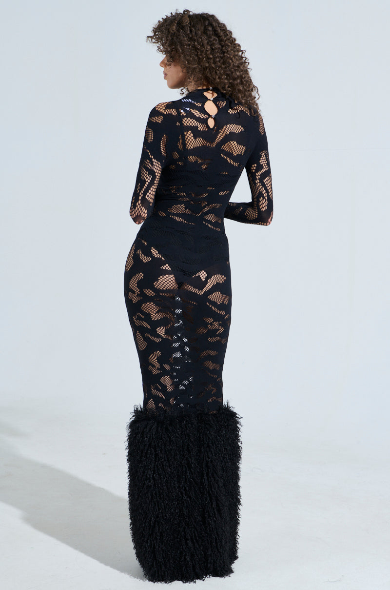 A person with curly hair, facing away, wears the ALL EYES ON ME SEAMLESS MAXI DRESS WITH FUR. The black dress features a seamless mesh design and a fluffy textured section from the knees down, exuding sophistication against a plain white background.
