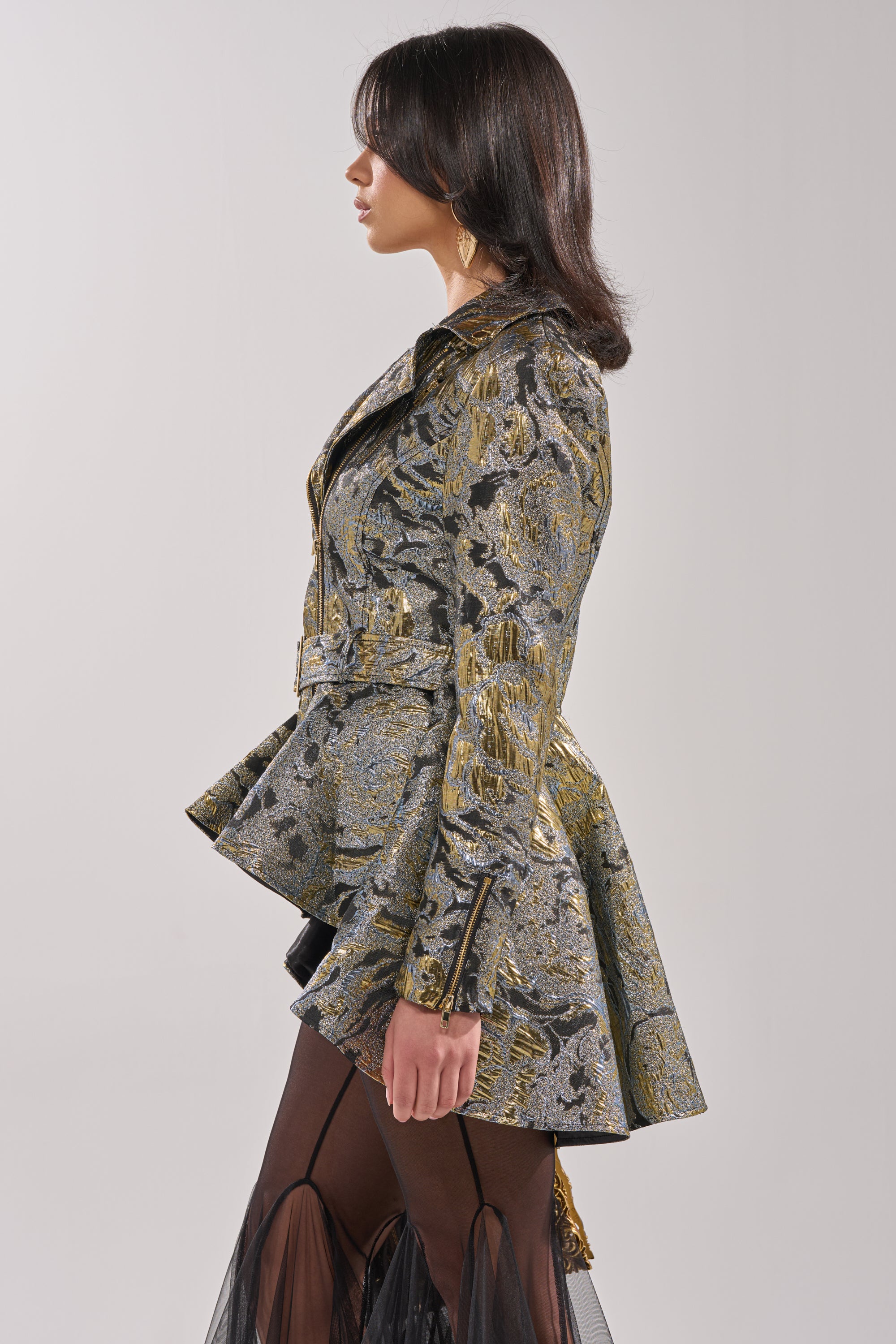 A woman with medium-length dark hair and large earrings stands in profile wearing the BROCADE HIGH LOW MOTO jacket, featuring a flared peplum and belted waist, paired with sheer black pants. 1
