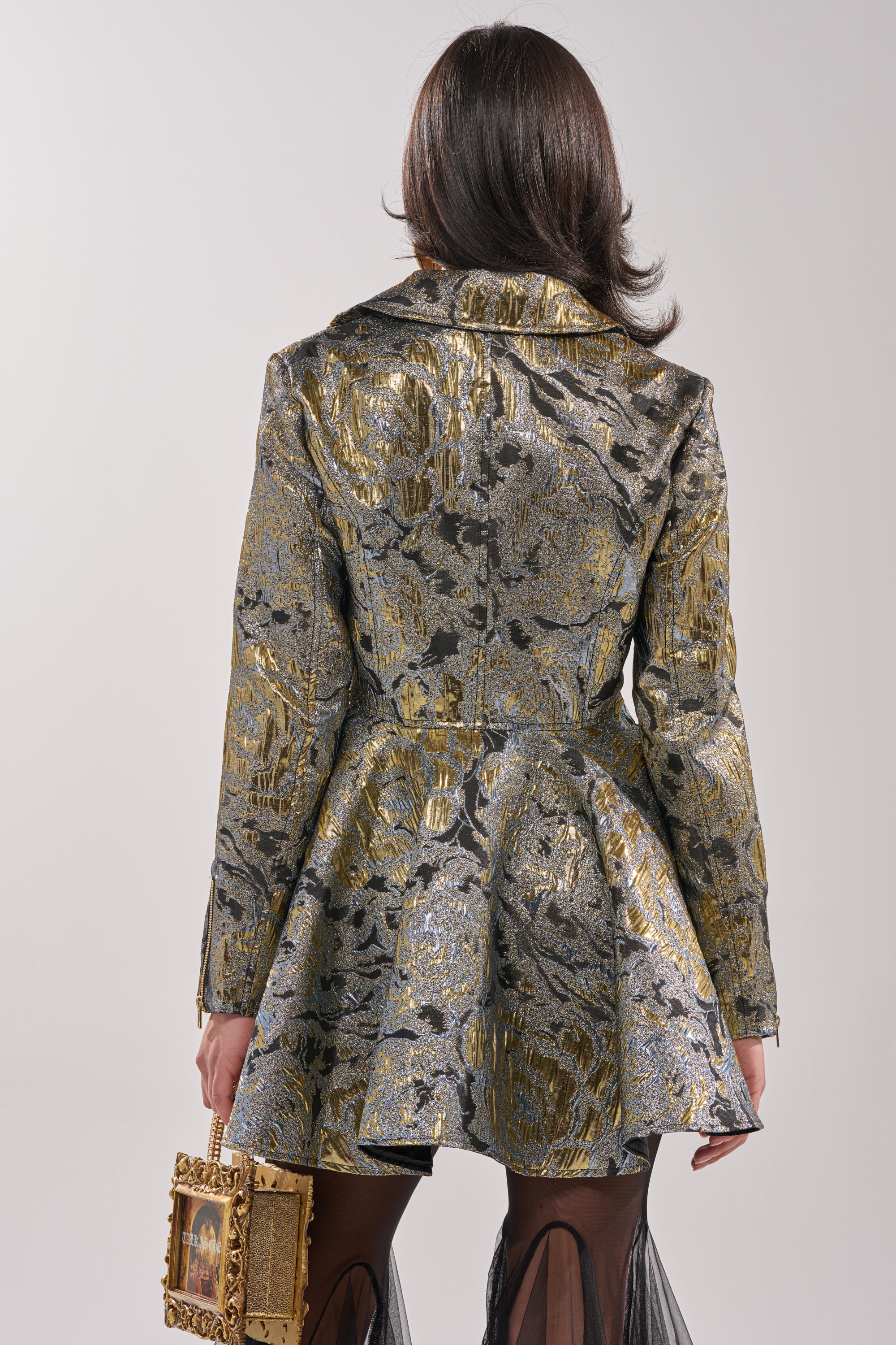 A person with shoulder-length dark hair, seen from behind, wears the BROCADE HIGH LOW MOTO jacket featuring a flared peplum hem and gold-and-silver floral patterns. They carry a small gold frame-shaped handbag and wear sheer black stockings. 2