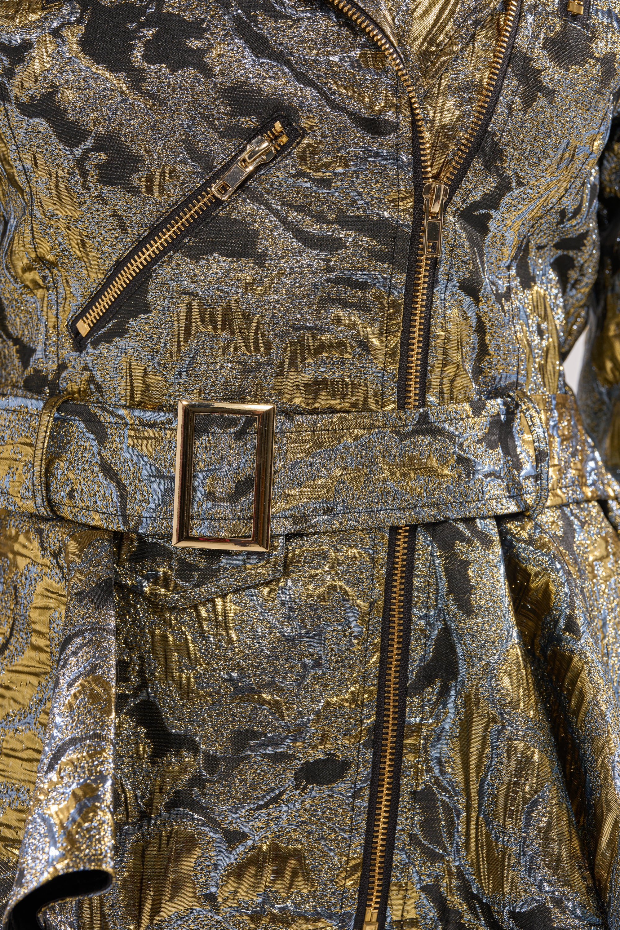 Close-up of the BROCADE HIGH LOW MOTO jacket, featuring a metallic brocade fabric, belted waist, zippered pocket and closure. The shimmering abstract pattern and high-low peplum hem make it a standout statement piece. 4