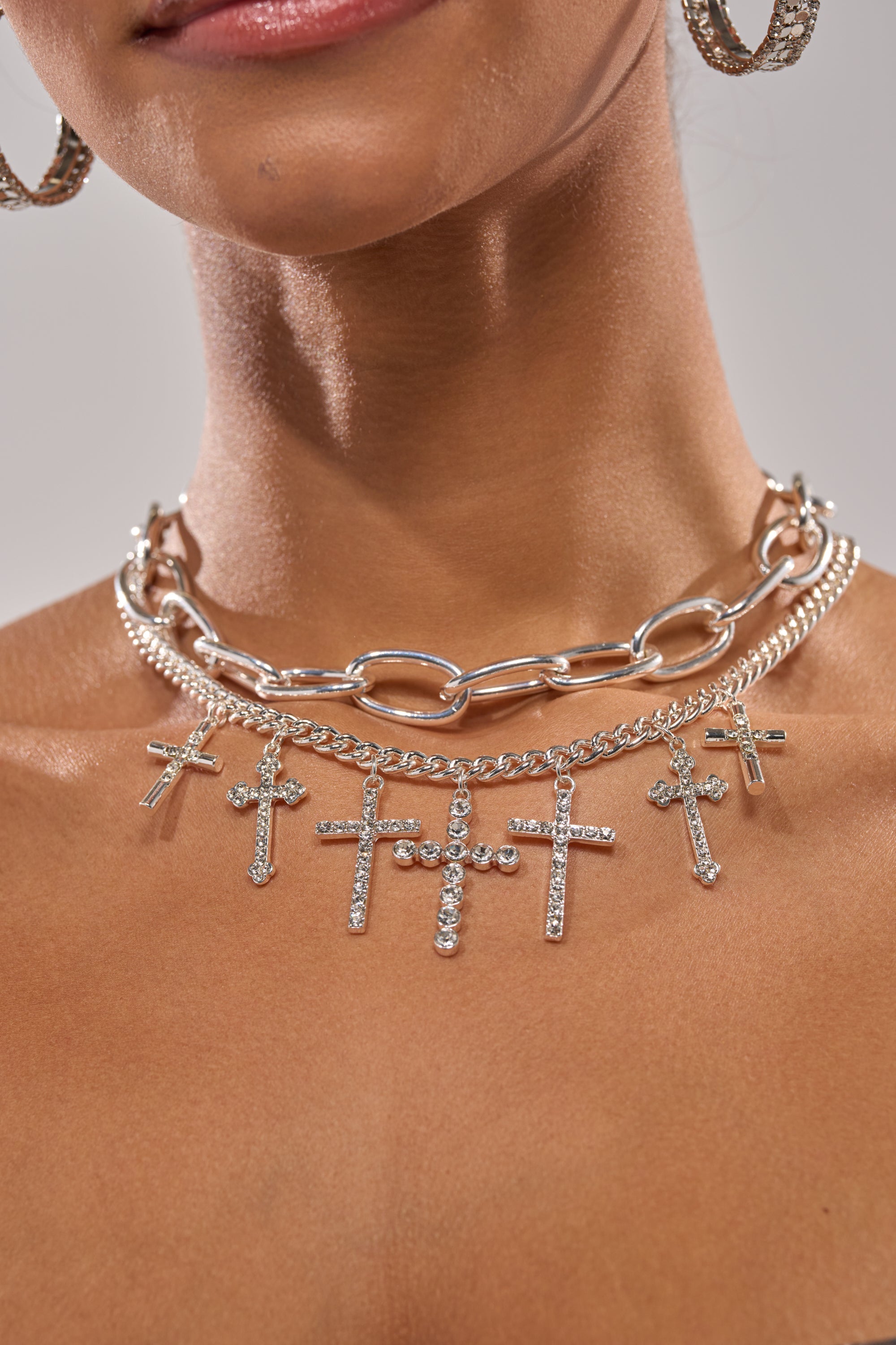 A close-up shows a woman wearing the CROSS MY HEART SILVER NECKLACE, featuring chunky chains and dazzling rhinestone cross pendants. Only her lower face, neck, and shoulders are visible. 1
