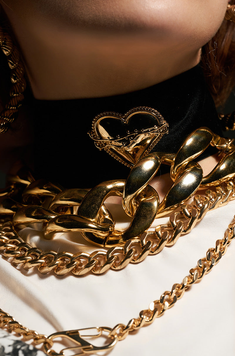 A close-up of a person wearing multiple gold chains around their neck showcases a faux gold heart pendant. The backdrop—a black and white shirt—enhances the contrast with the gleaming jewelry, while the ITALIAN LOVE CHOKER adds an element of elegance.