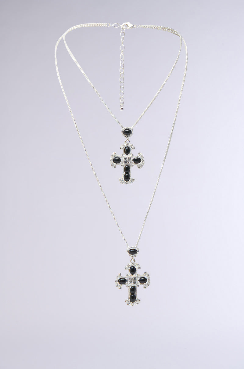 The IN MY PRAYERS SILVER NECKLACE showcases a layered design with two cross pendants adorned with onyx-like jewels; the shorter pendant sits above the longer one, creating a cascading effect against any plain backdrop.