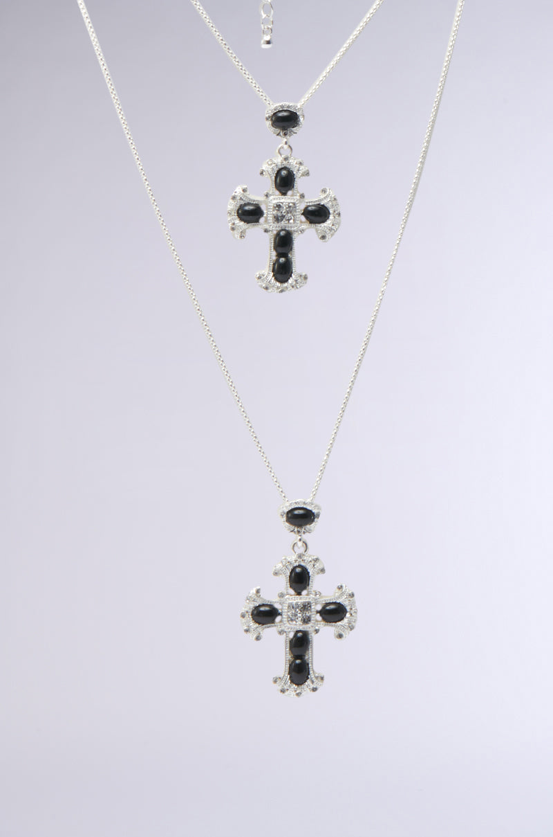 The IN MY PRAYERS SILVER NECKLACE features a cross-shaped pendant with onyx-inspired jewels and small clear gems, hanging on a thin chain against a plain background.