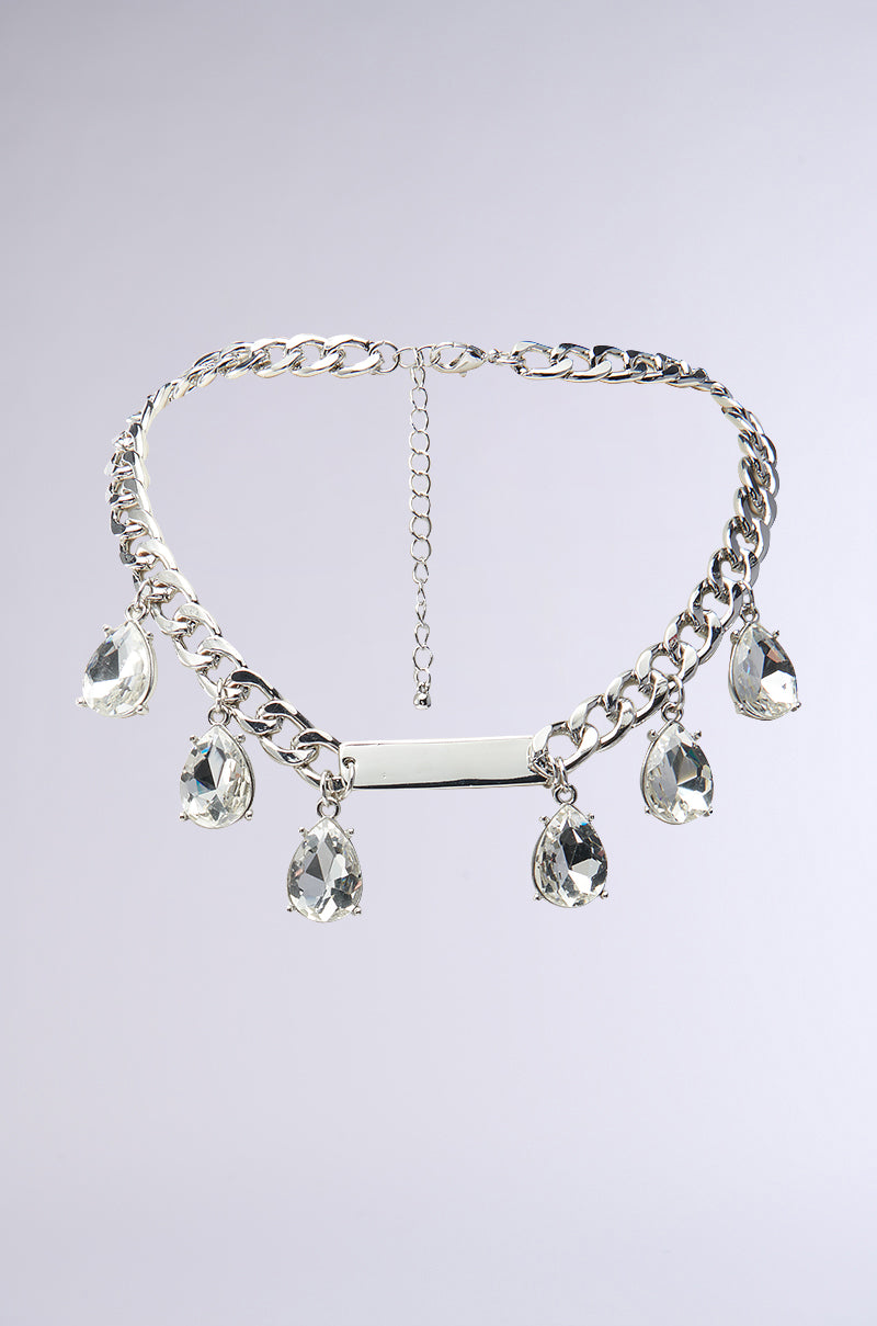 The STAY DRIPPING IN DIAMONDS NECKLACE features a silver curb chain, a central metal plate, and five large teardrop-shaped faux diamond pendants, all elegantly displayed on a light gray background.