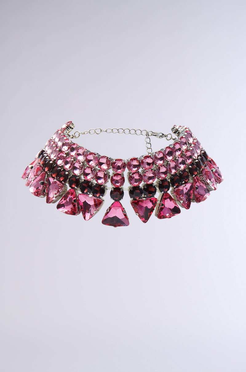 The SUCH A GEM STATEMENT CHOKER highlights vibrant pink gemstones in triangular and circular configurations, forming a shimmering display. It is equipped with an adjustable lobster clasp set against a simple gray backdrop.