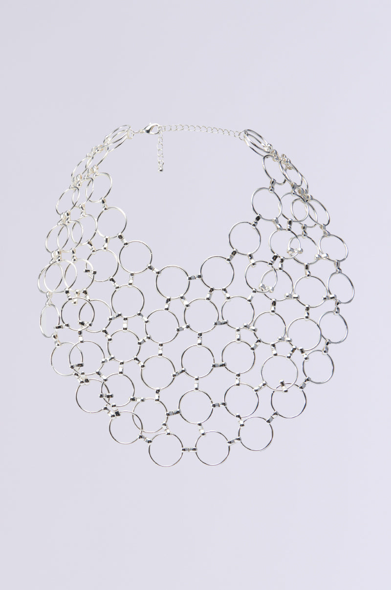 The RUNNING IN CIRCLES NECKLACE exhibits a stunning silver design with o-ring hardware, displaying interconnected circular links in a broad, net-like pattern. This eye-catching accessory includes a clasp and an adjustable chain, elegantly presented against a simple white backdrop.
