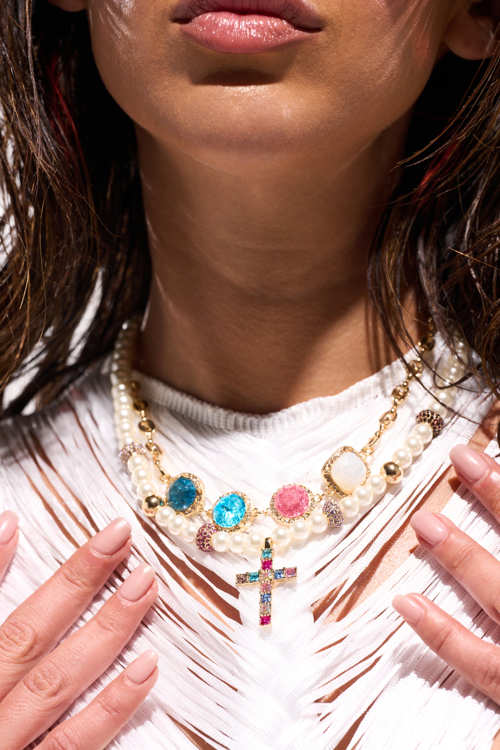 A close-up of a woman wearing the REGENCY NECKLACE, a statement pearl piece with colorful gemstones and a jeweled cross pendant, paired with a white textured top and manicured nails visible. 0