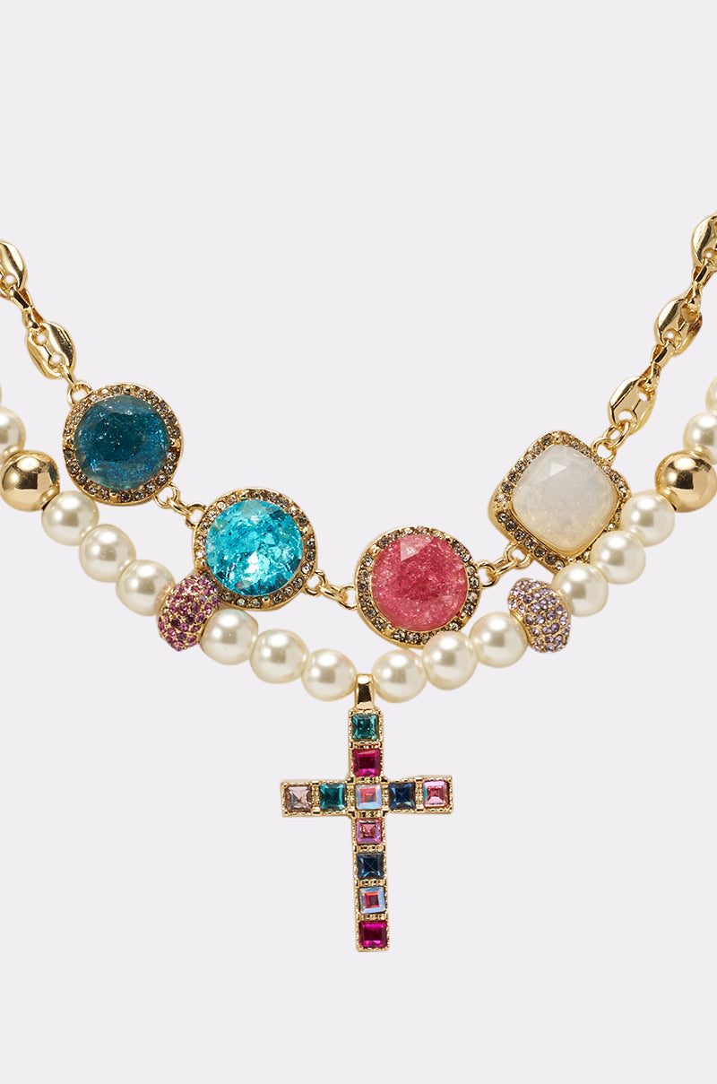 The REGENCY NECKLACE is a bold statement piece with a gold chain, colorful gemstones, layered white pearls, and a cross pendant embellished with multicolored gems. 2