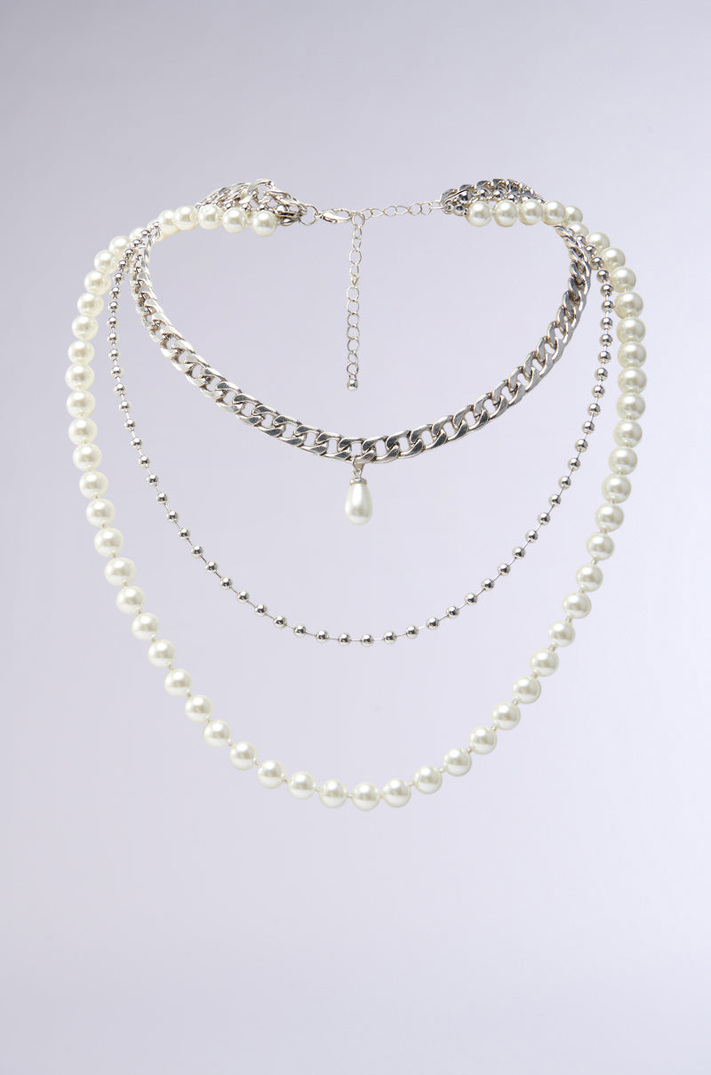 The GLAM LIFE LAYERED NECKLACE dazzles with its multi-layered faux silver design, featuring various chains and a pearl strand. An elegant pearl pendant on one chain adds contemporary flair against a neutral backdrop. 0