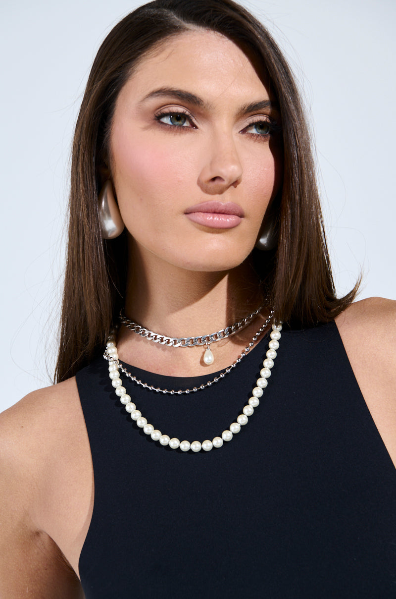 A woman with long brown hair wears a black sleeveless top and the stunning GLAM LIFE LAYERED NECKLACE featuring pearls and chains. Her pearl earrings complement her soft pink makeup, while a striking faux silver necklace adds an elegant touch against a plain white background. 1
