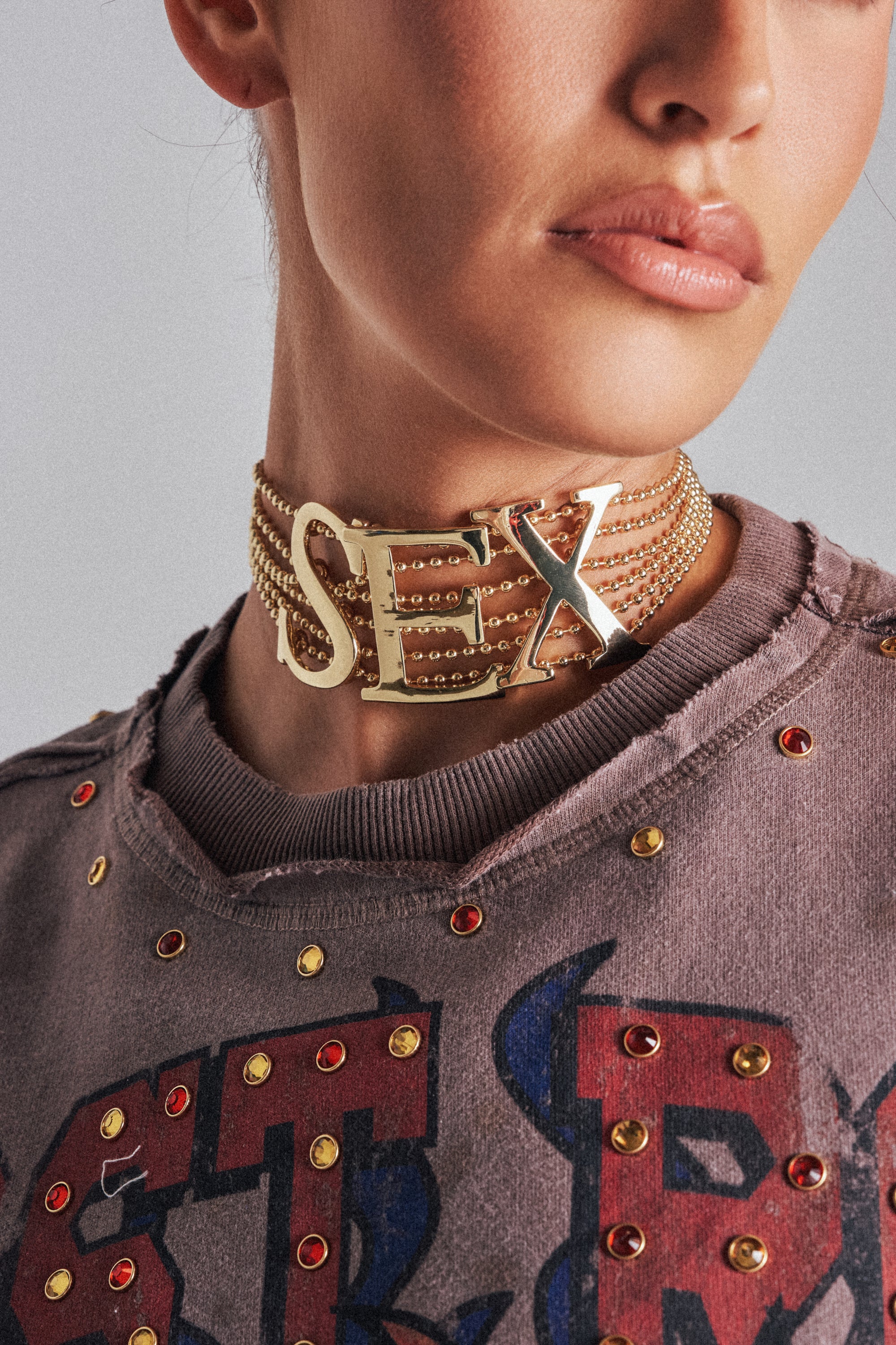 A person wears a distressed, studded shirt and the bold LETS DO IT NECKLACE featuring gold chains and large letter pendants. Only the lower face and neck are visible.