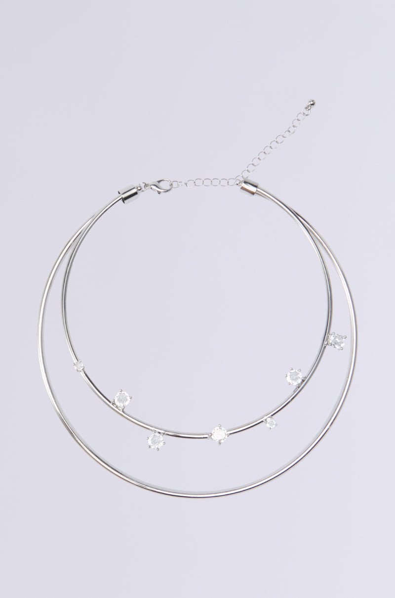 The FIND LESS NECKLACE is a striking statement piece featuring a silver double hoop design with rhinestone embellishments. The larger outer hoop elegantly encircles an inner one adorned with five small gemstones. It comes with an adjustable chain and clasp for secure fastening, showcased against a simple light gray background.