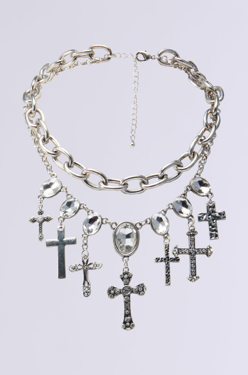The YOUR MUSE NECKLACE features a striking silver chain with large links, adorned with hanging cross pendants and oval gemstones. The adjustable clasp allows for customization, while the gemstone accents and diverse cross styles enhance its distinctive look.