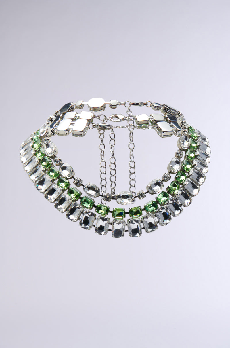 The BELLA LAYERED NECKLACE features a stunning faux silver design adorned with gemstone embellishments in green and clear shades, arranged in eye-catching geometric patterns. The necklace's chains and clasp elegantly rest at the back, all beautifully displayed against a light gray background.