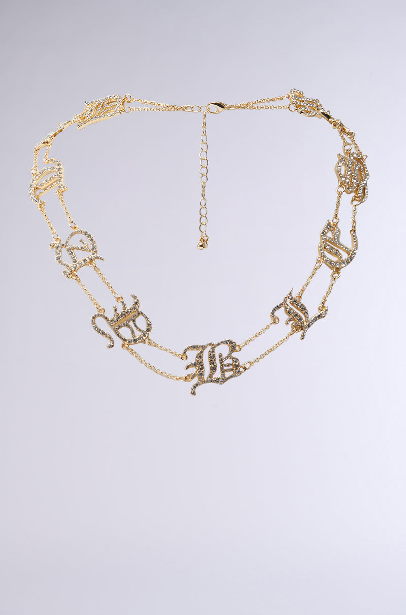 The STAY BLESSED NECKLACE is a faux gold piece featuring ornate, interlinked rhinestone letters encrusted with small diamonds. It has a dual-layer adjustable chain with a dangling clasp, set against a neutral background.