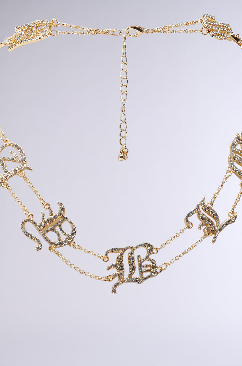 The STAY BLESSED NECKLACE features gold-toned double-layer chains adorned with ornate rhinestone letters encrusted with small crystals and a delicate adjustable clasp, set against a soft gradient gray background.