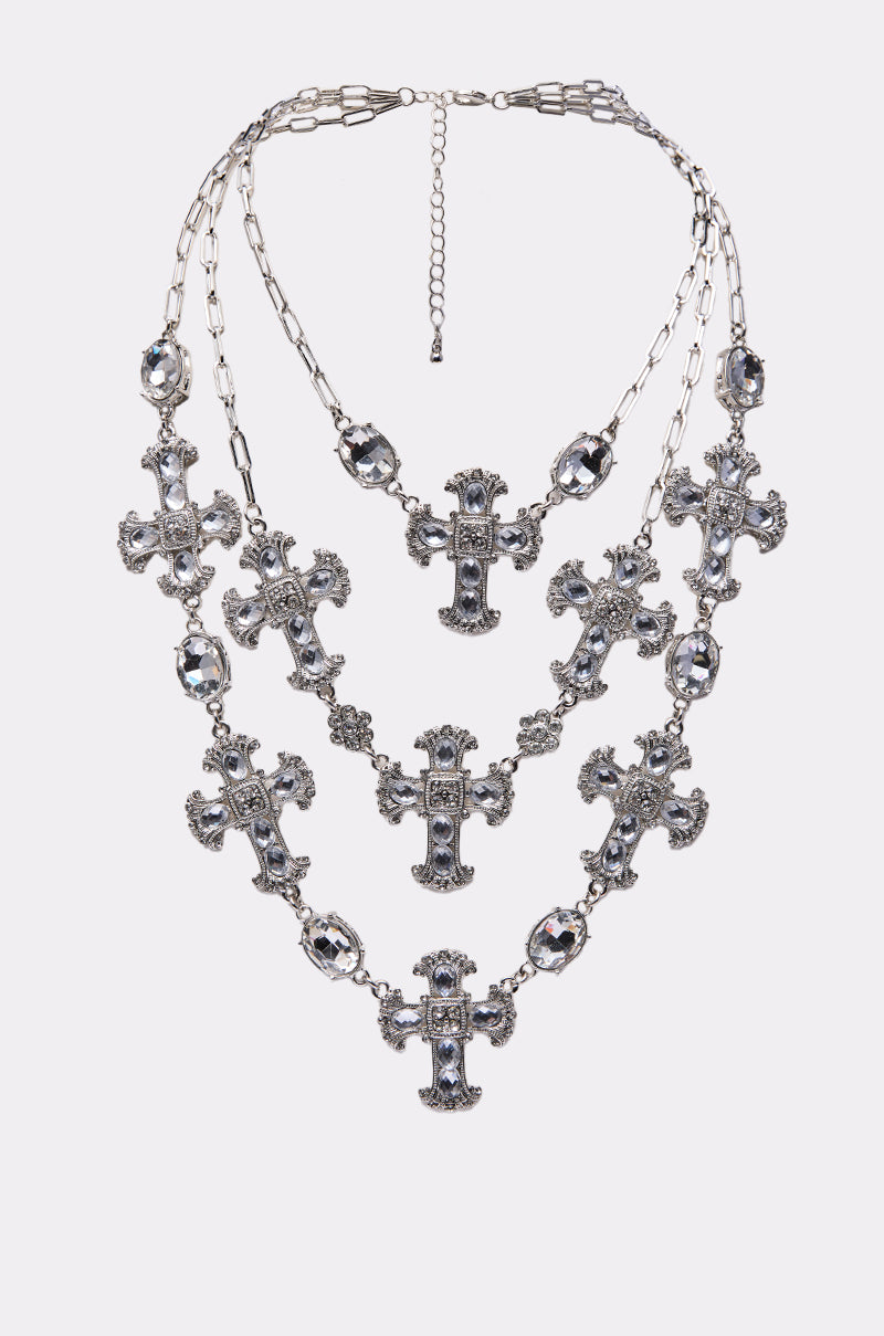 The SO TRENDY NECKLACE is a layered chain with ornate cross charms and oval gemstones, featuring intricate details and adjustable links, shown on a plain light background.