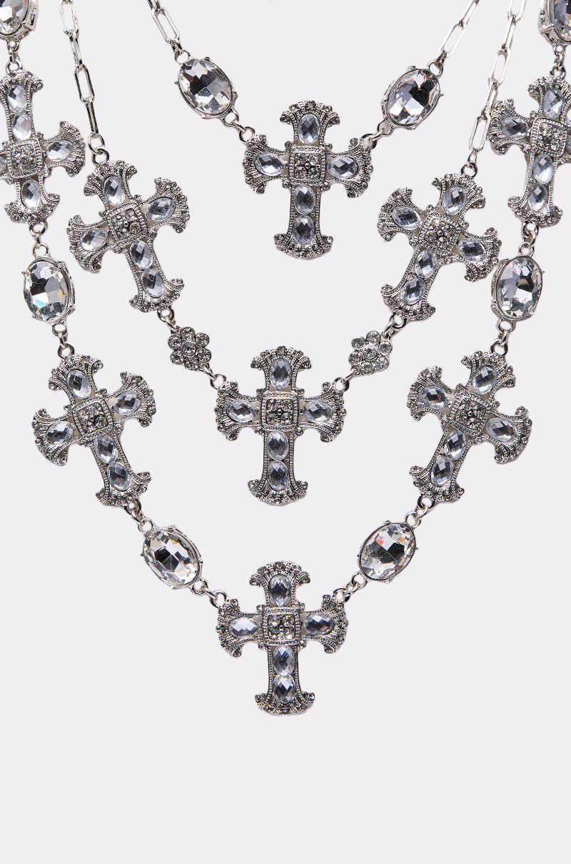The SO TRENDY NECKLACE showcases layered chains with ornate cross charms and faceted oval crystals, elegantly arranged against a white background.