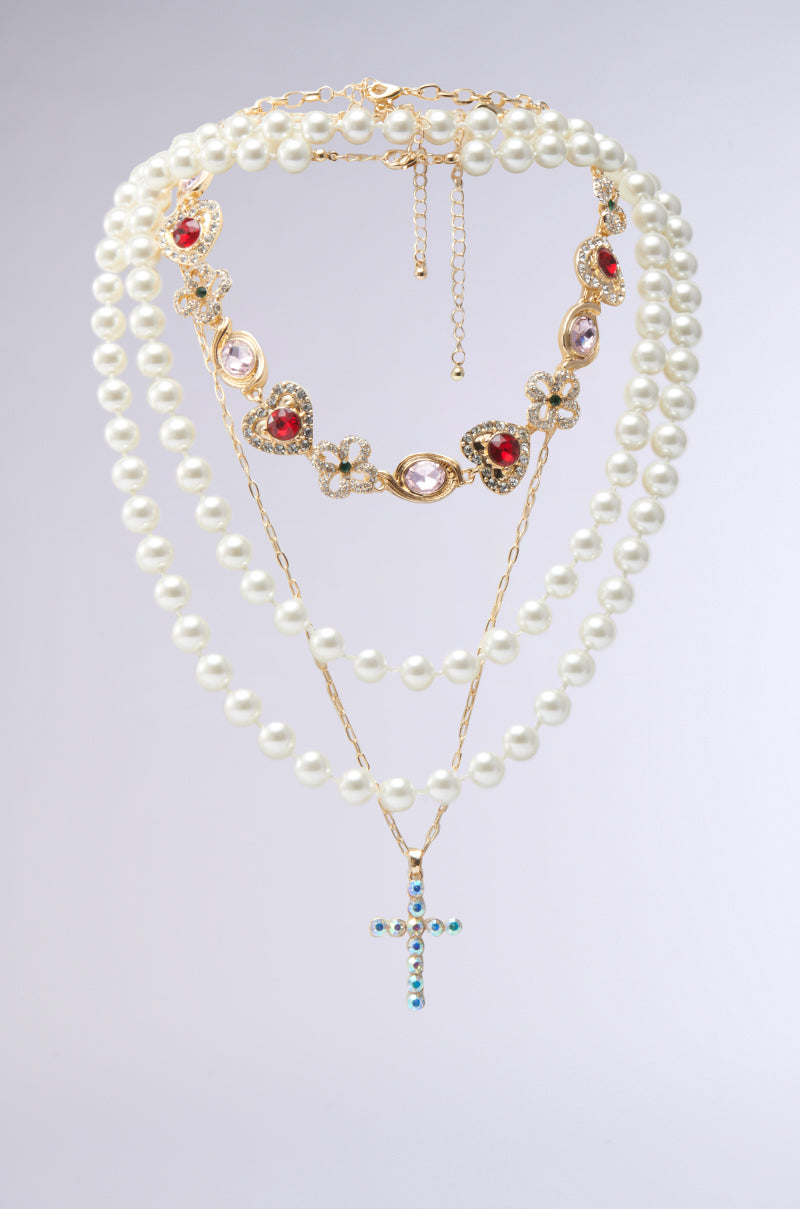 Introducing the RENAISSANCE VIBES NECKLACE: a set of three layered necklaces featuring oversized pearls and gold-toned chains. The shortest has red and purple jewels, the middle is a simple pearl strand, and the longest features a blue-jeweled cross pendant. 0