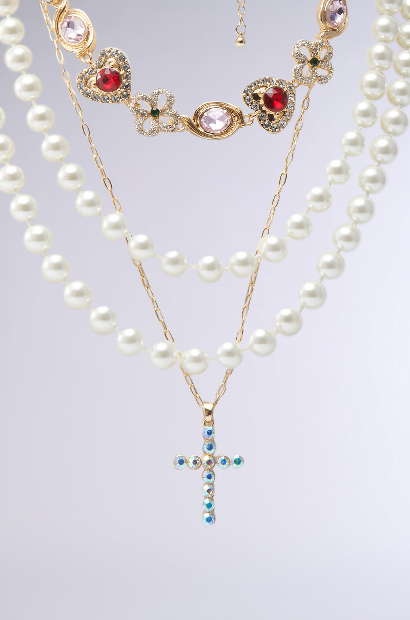 The RENAISSANCE VIBES NECKLACE Set features layered necklaces with oversized pearls, a gold chain adorned with heart-shaped and gemstone details, and a cross pendant embellished with blue and clear stones on a light gray background. 1