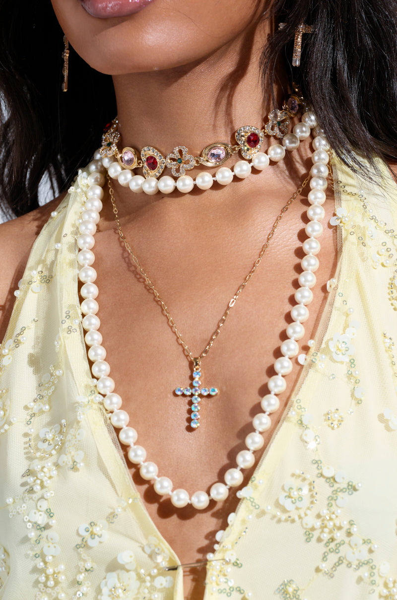 A woman models a layered necklace look featuring the RENAISSANCE VIBES NECKLACE with oversized pearls, a pearl choker embellished with colorful gems, and a gold chain displaying a blue gem cross pendant. She pairs them with her light yellow, floral dress and gold-toned dangling earrings. 2